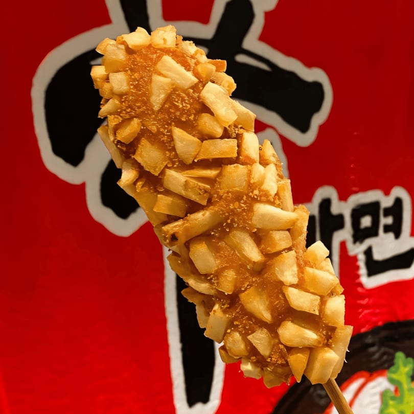 A Korean corn dog, coated in crispy french fries, against a red and black background.