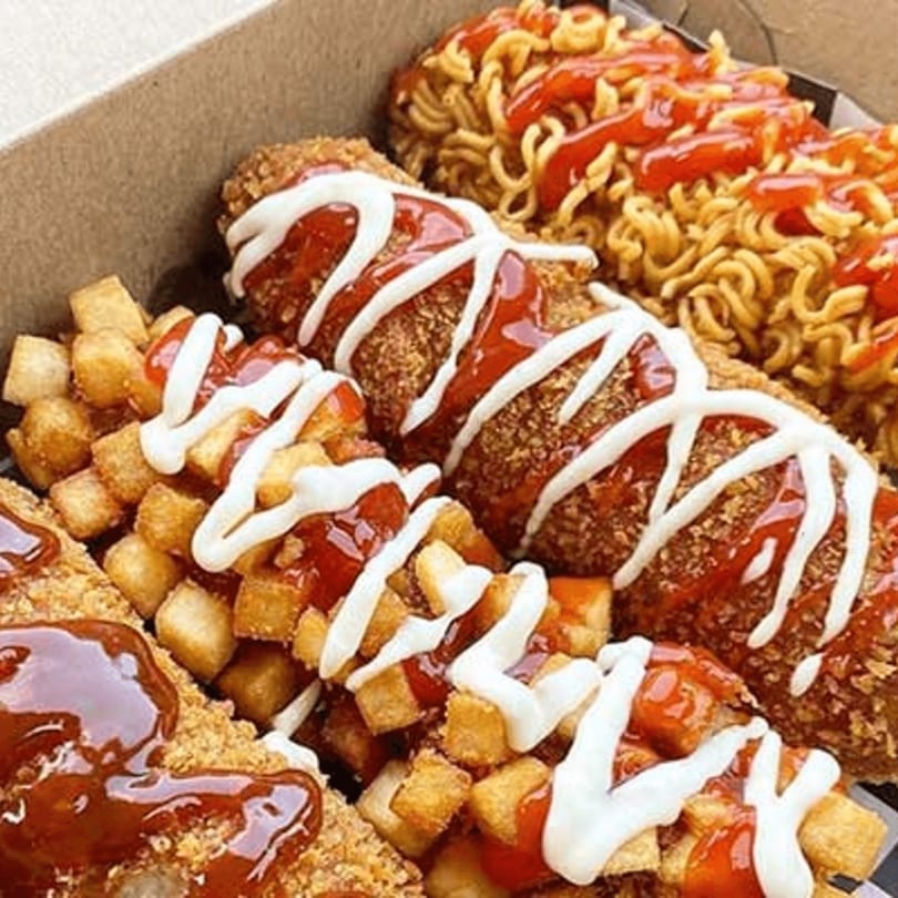 Korean corn dogs with various toppings, including ramen, tater tots, ketchup, and white sauce.